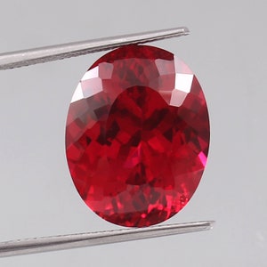 May include: A faceted, oval-shaped, deep red gemstone is held by a pair of silver tweezers. The gem has a brilliant, reflective surface, showcasing its many facets. The gemstone is a vibrant, saturated red color.