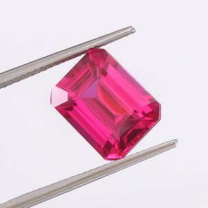 Könnte beinhalten: Ein leuchtend rosa Edelstein mit Smaragdschliff, gehalten von einer Pinzette vor weißem Hintergrund.
