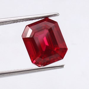 Puede incluir: Una piedra preciosa de color rojo oscuro tallada en esmeralda, sostenida por unas pinzas sobre un fondo blanco.