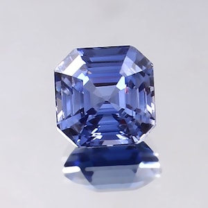 May include: A square-shaped, faceted sapphire gemstone with a deep blue colour. The gemstone is cut in a step-cut style, creating a series of flat facets that reflect light.