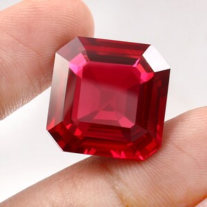 May include: A square-cut, deep red gemstone with a faceted surface that reflects light. The stone features sharp, clean edges and a transparent, vibrant crimson color, showcasing a symmetrical geometric design with multiple polished facets.