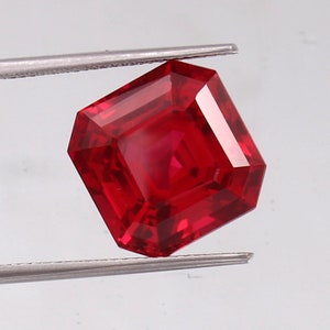 May include: A faceted, rectangular-cut ruby gemstone held by tweezers. The gem is a deep, saturated red color with sharp, clean edges and a high degree of clarity. The gemstone is displayed against a light background.