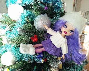 Può includere: Una bambola con capelli viola e un cappello di pelliccia bianco, indossa un maglione bianco e una gonna viola, è posizionata su un albero di Natale decorato. L'albero è adornato con ornamenti argentati, orpelli blu e bacche rosse, creando una scena festosa.