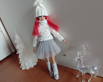 Può includere: Una bambola con i capelli rossi che indossa un cappello di lana bianco, un maglione bianco e una gonna di tulle argentata. La bambola è in piedi su un pavimento di legno con un albero di Natale bianco scintillante e una statuetta di renna di plastica trasparente.