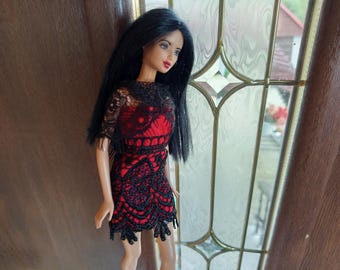 May include: A doll wearing a red and black lace dress with black hair. The dress has a fitted bodice and a flared skirt, with intricate lace detailing. The doll is standing near a window with a decorative glass design.