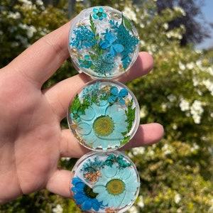 May include: Three clear, round phone grips with embedded dried flowers in shades of blue and gold. The top grip features small blue flowers and gold flakes. The middle grip has a light blue daisy design. The bottom grip has a blue daisy design.
