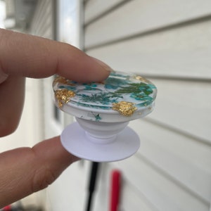 Pu&ograve; includere: Un supporto per telefono bianco con una parte superiore trasparente, incastonato con fiori secchi e accenti di foglie d'oro. L'impugnatura ha una parte superiore rotonda e una base bianca. Il design presenta elementi floreali turchese e bianchi.
