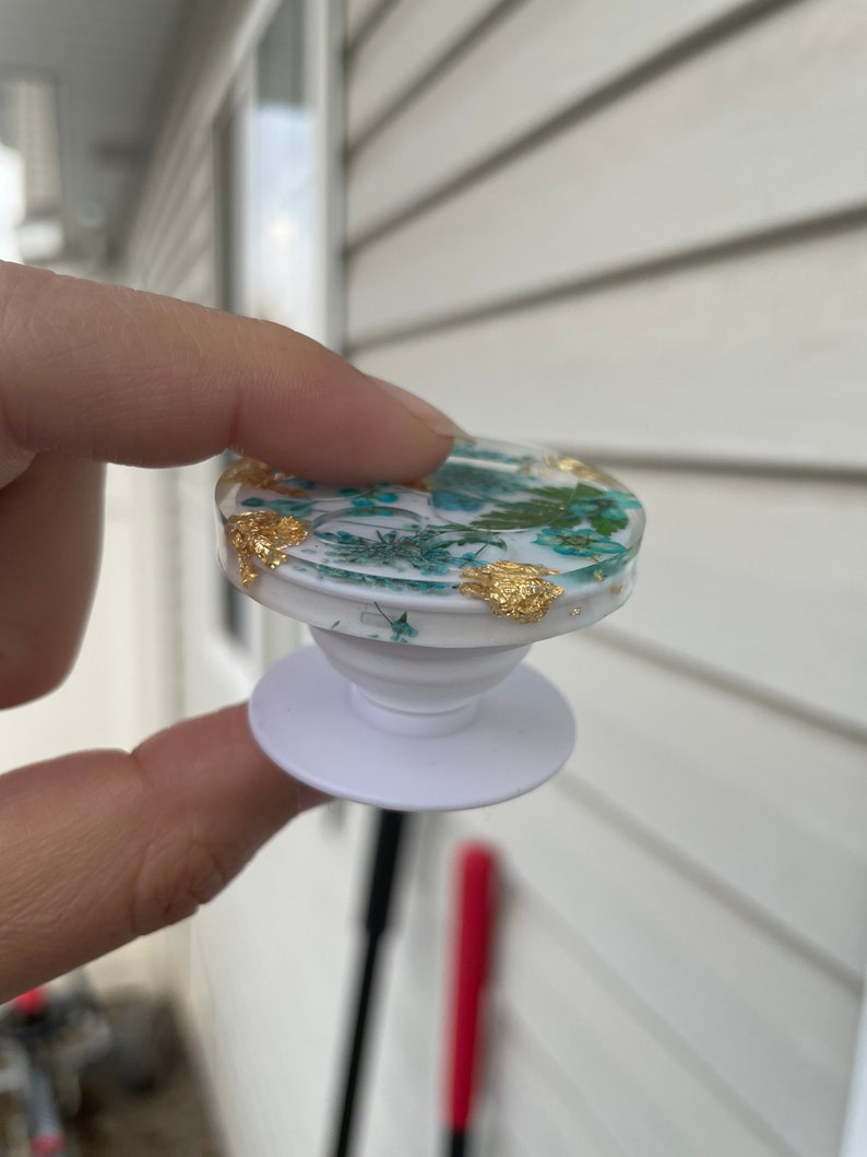 Pu&ograve; includere: Un supporto per telefono bianco con una parte superiore trasparente, incastonato con fiori secchi e accenti di foglie d'oro. L'impugnatura ha una parte superiore rotonda e una base bianca. Il design presenta elementi floreali turchese e bianchi.
