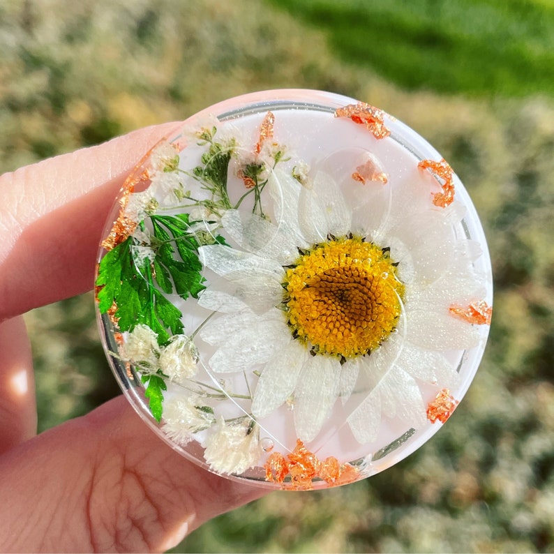 Pu&ograve; includere: Un sottobicchiere rotondo e trasparente in resina con una grande margherita bianca e verde secco. Il sottobicchiere ha accenti di foglia d'oro e uno sfondo rosa chiaro. Un oggetto di arredamento floreale unico e fatto a mano.