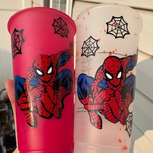 May include: Two reusable cups with a Spiderman design. One cup is pink with a white spider web design and the other is white with orange splatter paint and a black spider web design. Both cups feature a cartoon image of Spiderman.