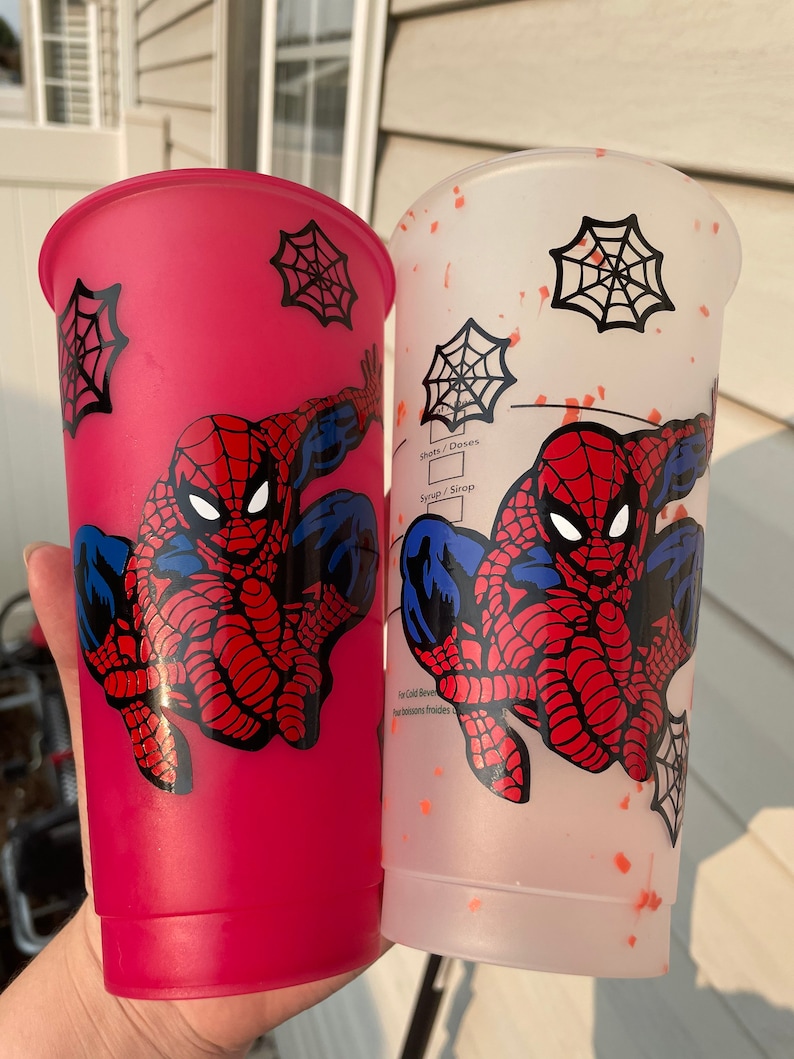 May include: Two reusable cups with a Spiderman design. One cup is pink with a white spider web design and the other is white with orange splatter paint and a black spider web design. Both cups feature a cartoon image of Spiderman.