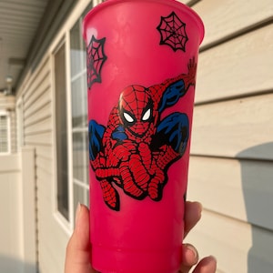 May include: A pink reusable cup with a Spiderman design. The design features Spiderman in a red and blue suit with a black spider web pattern. There are also black spider webs on the cup.