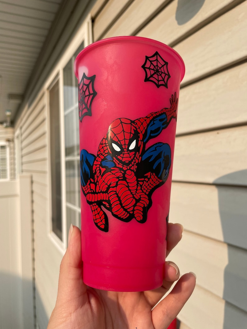 May include: A pink reusable cup with a Spiderman design. The design features Spiderman in a red and blue suit with a black spider web pattern. There are also black spider webs on the cup.