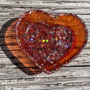 Könnte beinhalten: Ein herzförmiges Schmucktablett mit einem transparenten, bernsteinfarbenen Äußeren und einem mit rotem Glitzer gefüllten Inneren. Die Schale hat einen erhöhten Rand mit silbernen Glitzerakzenten. Das Tablett liegt auf einer verwitterten Holzoberfläche.