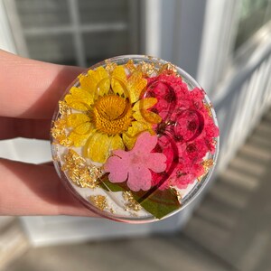 Pu&ograve; includere: Un supporto per telefono rotondo e trasparente con fiori secchi gialli, rosa e rossi, impreziosito da scaglie d'oro. La composizione floreale &egrave; incastonata in resina trasparente, creando un accessorio decorativo e funzionale.
