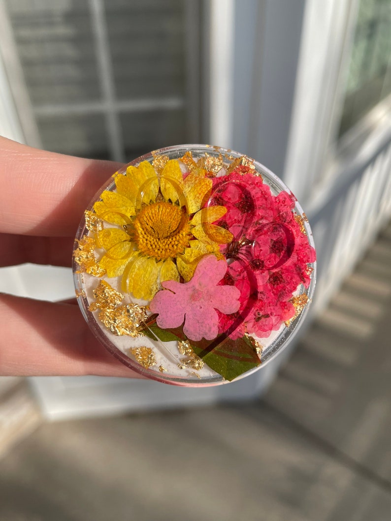 Pu&ograve; includere: Un supporto per telefono rotondo e trasparente con fiori secchi gialli, rosa e rossi, impreziosito da scaglie d'oro. La composizione floreale &egrave; incastonata in resina trasparente, creando un accessorio decorativo e funzionale.