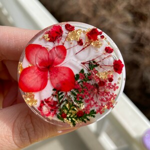 Pu&ograve; includere: Un oggetto rotondo e trasparente in resina con fiori rossi, scaglie di foglie d'oro e fogliame verde incorporati. Il design presenta un grande fiore rosso al centro, circondato da fiori rossi pi&ugrave; piccoli e accenti dorati. L'oggetto &egrave; tenuto in mano.