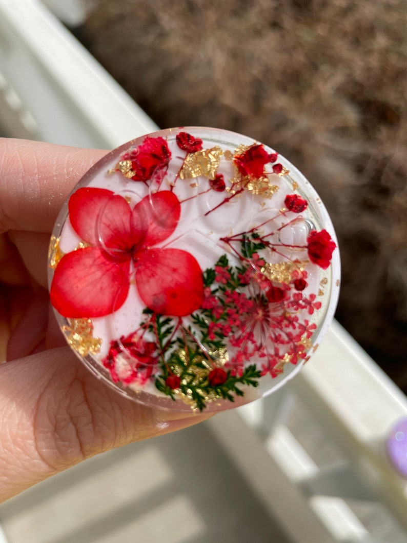 Pu&ograve; includere: Un oggetto rotondo e trasparente in resina con fiori rossi, scaglie di foglie d'oro e fogliame verde incorporati. Il design presenta un grande fiore rosso al centro, circondato da fiori rossi pi&ugrave; piccoli e accenti dorati. L'oggetto &egrave; tenuto in mano.