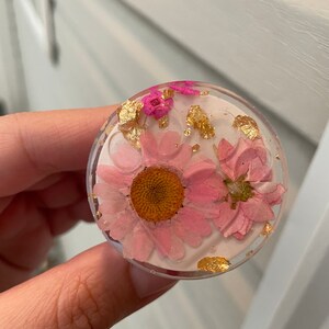 Pu&ograve; includere: Un supporto per telefono rotondo e trasparente con fiori rosa secchi e inclusioni di foglie d'oro in resina. Il supporto ha uno sfondo rosa chiaro e un bordo trasparente. I fiori includono una margherita e fiori pi&ugrave; piccoli.