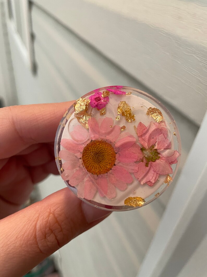 Pu&ograve; includere: Un supporto per telefono rotondo e trasparente con fiori rosa secchi e inclusioni di foglie d'oro in resina. Il supporto ha uno sfondo rosa chiaro e un bordo trasparente. I fiori includono una margherita e fiori pi&ugrave; piccoli.