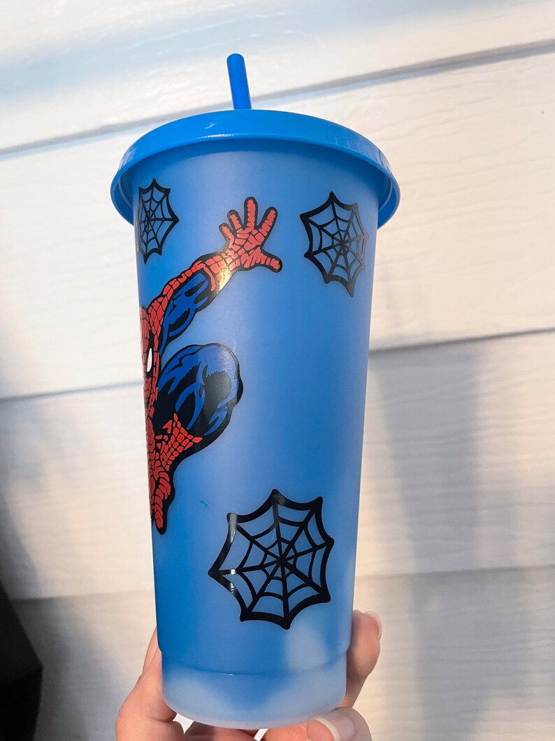 May include: A blue reusable cup with a Spider-Man design and a red straw. The cup has a white swirl design at the bottom and four black spiderwebs.