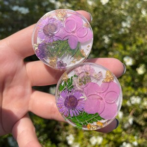 Pu&ograve; includere: Due supporti rotondi e trasparenti per telefono con fiori secchi e accenti di foglie d'oro. Ogni supporto presenta un fiore viola, un fiore rosa e foglie verdi racchiuse in resina. I supporti sono tenuti contro uno sfondo verde sfocato.