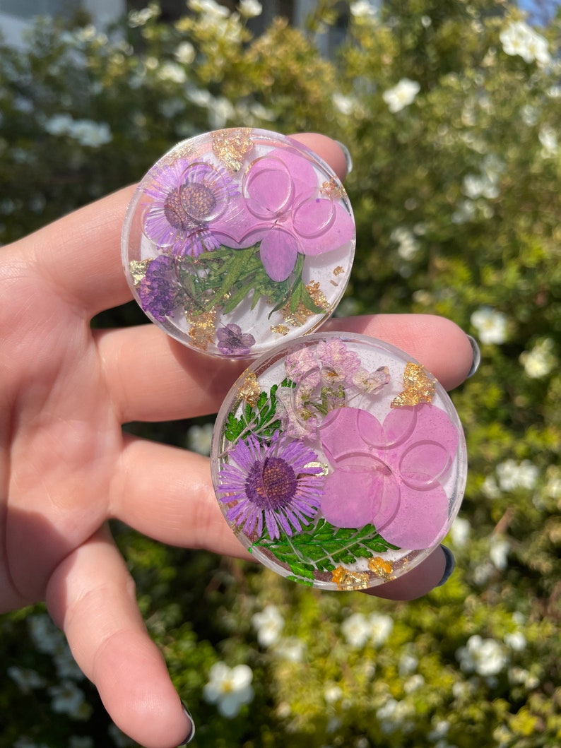 Pu&ograve; includere: Due supporti rotondi e trasparenti per telefono con fiori secchi e accenti di foglie d'oro. Ogni supporto presenta un fiore viola, un fiore rosa e foglie verdi racchiuse in resina. I supporti sono tenuti contro uno sfondo verde sfocato.