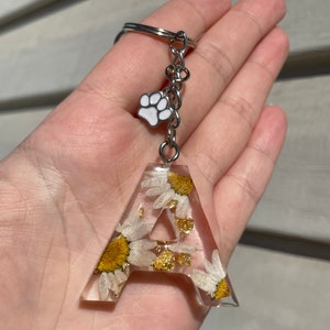 Può includere: Portachiavi con lettera "A" in resina trasparente con margherite essiccate e scaglie d'oro. Una catena d'argento tiene la lettera e un ciondolo con un'impronta di zampa bianca.