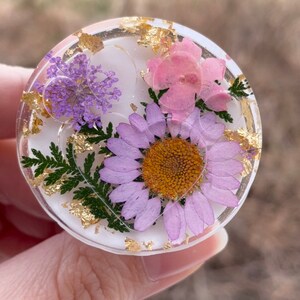 Pu&ograve; includere: Un oggetto rotondo in resina trasparente con fiori secchi e scaglie d'oro incorporate. Contiene una margherita viola, fiori rosa e rametti viola. La resina &egrave; posta su uno sfondo bianco.