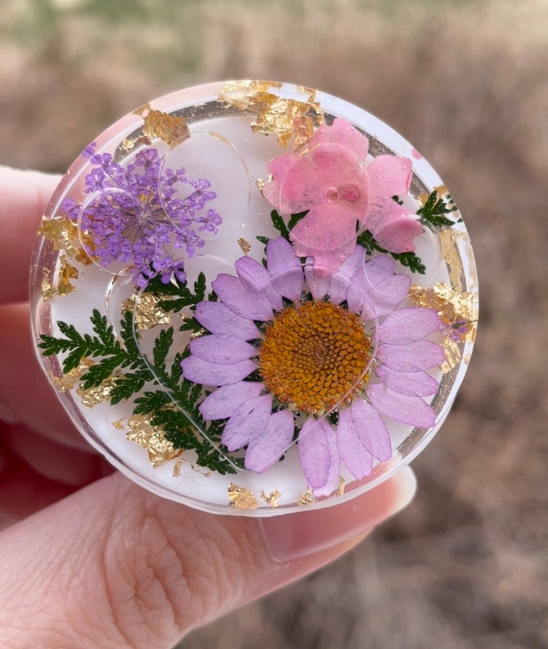 Pu&ograve; includere: Un oggetto rotondo in resina trasparente con fiori secchi e scaglie d'oro incorporate. Contiene una margherita viola, fiori rosa e rametti viola. La resina &egrave; posta su uno sfondo bianco.