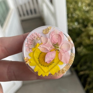 Pu&ograve; includere: Un supporto per telefono rotondo e trasparente con un design floreale. Presenta un girasole giallo, fiori rosa e accenti di foglie d'oro racchiusi in resina. Lo sfondo &egrave; bianco.