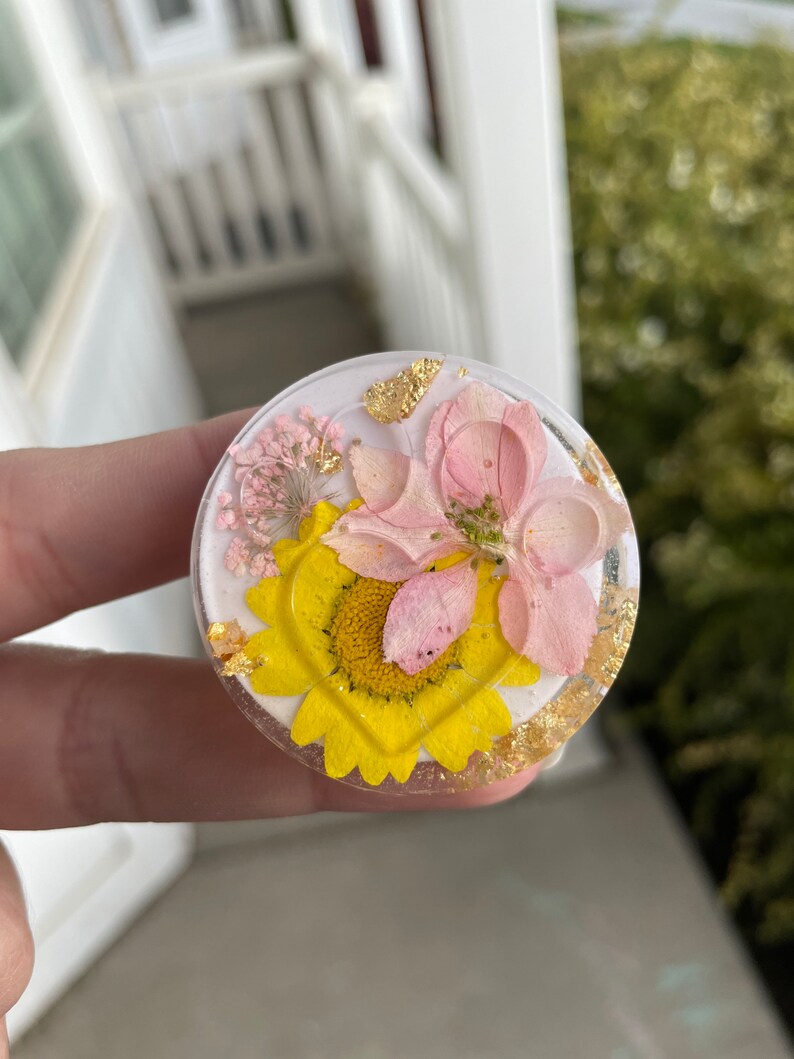 Pu&ograve; includere: Un supporto per telefono rotondo e trasparente con un design floreale. Presenta un girasole giallo, fiori rosa e accenti di foglie d'oro racchiusi in resina. Lo sfondo &egrave; bianco.