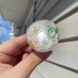 Pu&ograve; includere: Un supporto per telefono rotondo e trasparente con un design a zampa, contenente fiori secchi e scaglie d'oro. Il supporto &egrave; tenuto in mano su uno sfondo esterno sfocato.