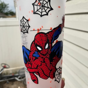 May include: A white reusable cup with a red and blue Spiderman design. The cup has a black spiderweb design and red splatter paint.