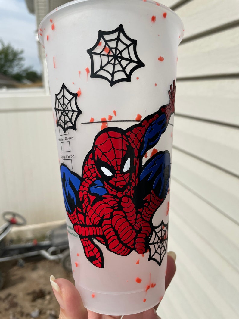 May include: A white reusable cup with a red and blue Spiderman design. The cup has a black spiderweb design and red splatter paint.