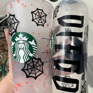 May include: A clear plastic water bottle with a green lid and a black spiderweb design. The bottle has the word "BOO" written on it in black letters. Next to the bottle is a clear plastic cup with a white and red speckled design and a black spiderweb design. The cup has the Starbucks logo on it.