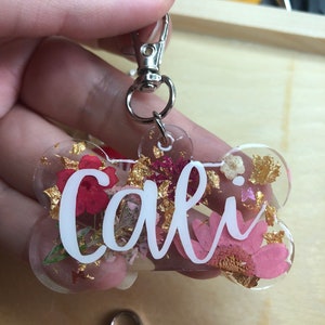Può includere: Un portachiavi trasparente a forma di osso con il nome "Cali" in corsivo bianco. Il portachiavi è riempito con fiori rosa e rossi secchi, scaglie d'oro e un fermaglio argentato.
