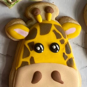 May include: A yellow and brown giraffe cookie with black eyes and a smiling mouth. The cookie is decorated with brown spots and has a light brown face.