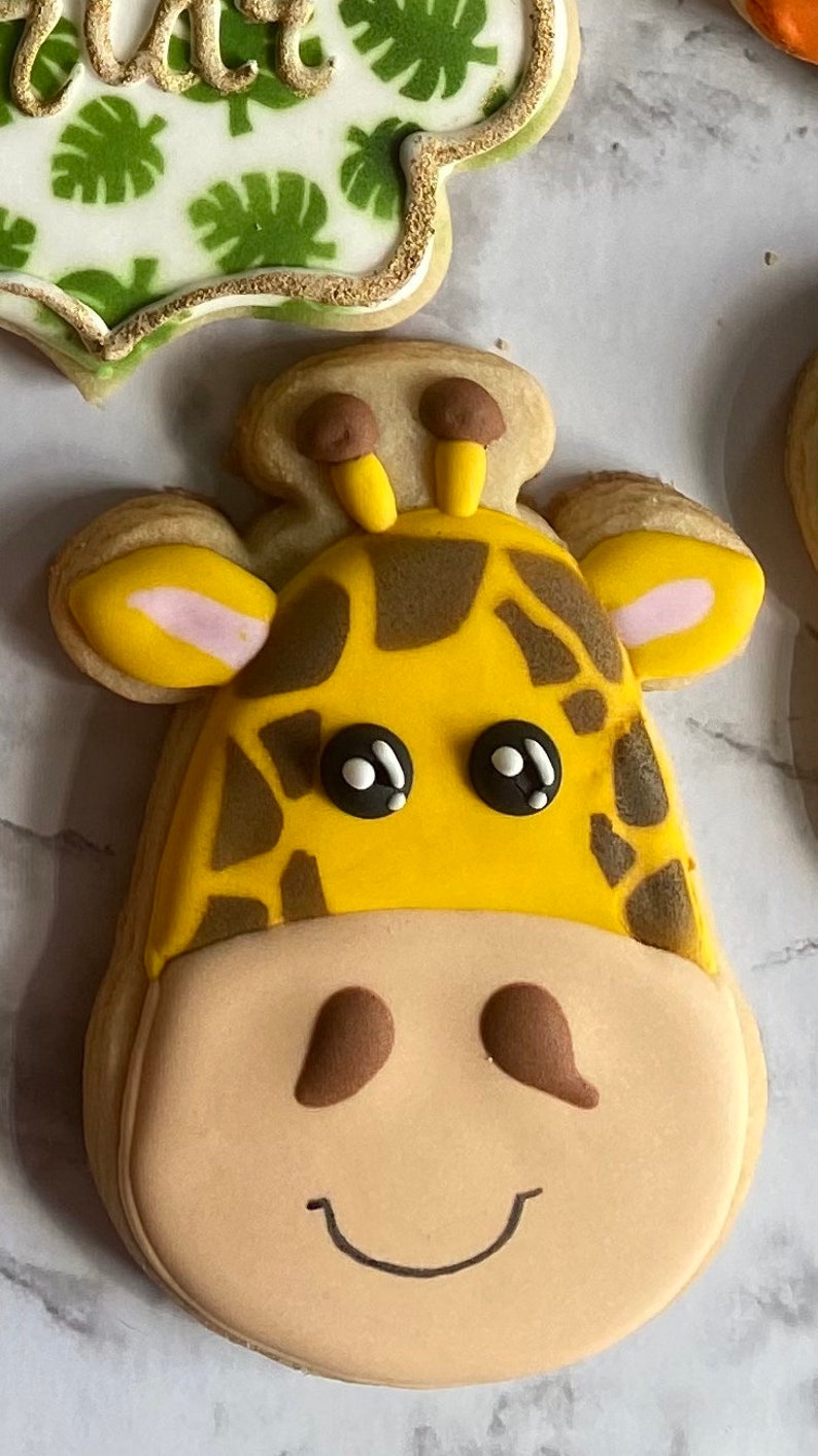 May include: A yellow and brown giraffe cookie with black eyes and a smiling mouth. The cookie is decorated with brown spots and has a light brown face.