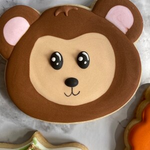 May include: A brown and tan cookie decorated with a monkey face. The monkey has black eyes, a black nose, and a small smile. The cookie is decorated with pink ears.