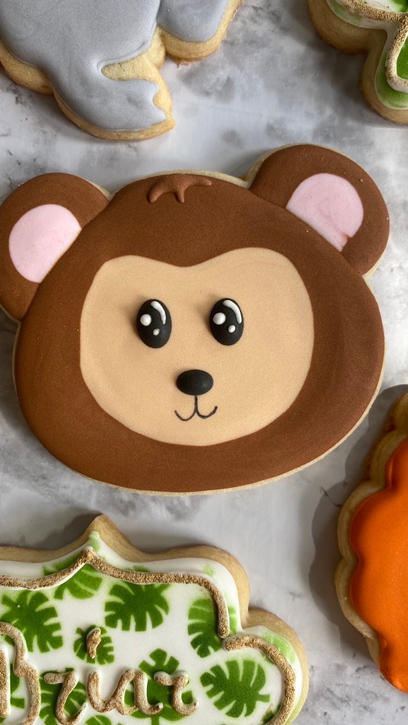 May include: A brown and tan cookie decorated with a monkey face. The monkey has black eyes, a black nose, and a small smile. The cookie is decorated with pink ears.