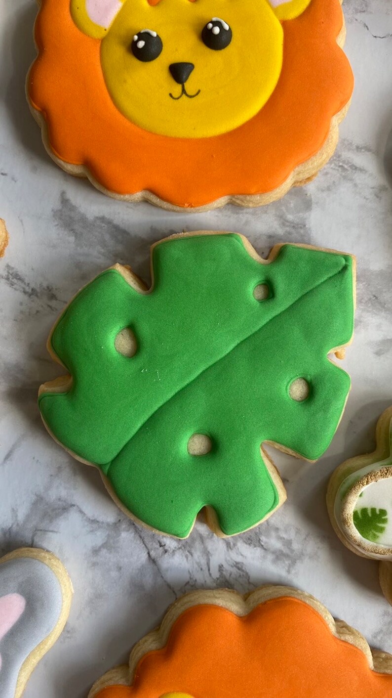 May include: A set of four decorated sugar cookies featuring a lion, a green leaf, and two other cookies. The cookies are decorated with colorful icing and have a whimsical, cartoon-like style.