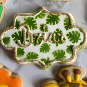 May include: A white cookie decorated with green leaves and gold glitter, with the name "Brix" written in gold icing.