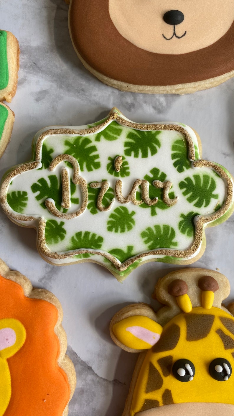 May include: A white cookie decorated with green leaves and gold glitter, with the name "Brix" written in gold icing.
