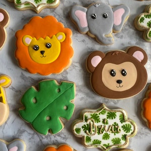 May include: A set of decorated sugar cookies featuring various jungle animals, including a lion, giraffe, monkey, and elephant. The cookies are decorated with colorful icing and have a whimsical, cartoon-like style. The cookies are arranged on a light gray marble surface. One cookie has the name "Brian" written on it.