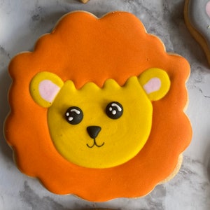 May include: A decorated sugar cookie with orange icing depicting a lion's face with yellow fur, black eyes, and a pink nose.