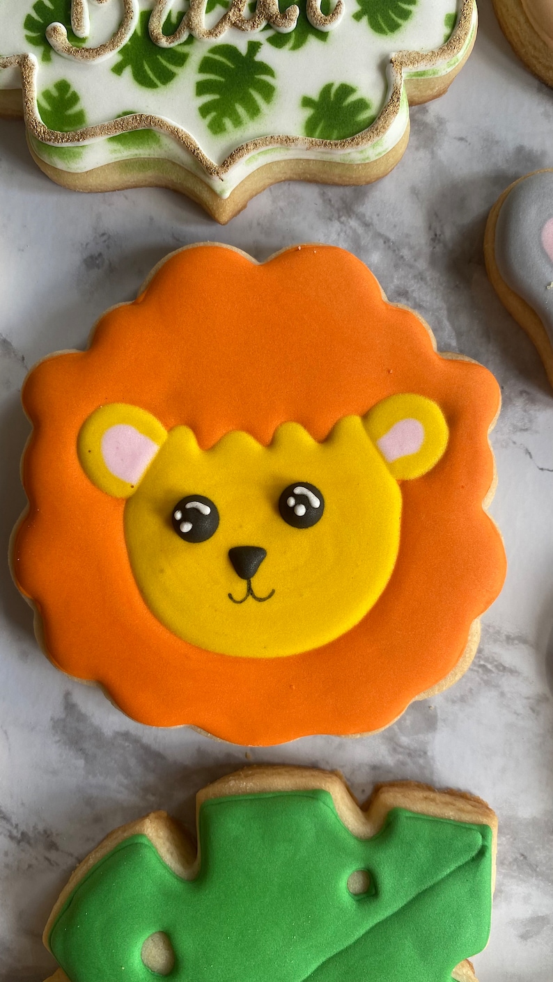 May include: A decorated sugar cookie with orange icing depicting a lion's face with yellow fur, black eyes, and a pink nose.
