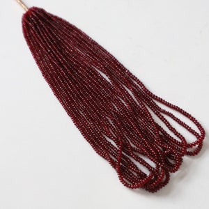 Può includere: Un filo di perle di rubino rosso scuro, lungo circa 38 cm, infilate su un filo sottile. Le perle sono piccole e di dimensioni uniformi, creando un aspetto liscio ed elegante.