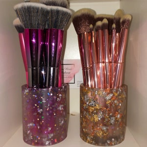 Puede incluir: Dos portacepillos de maquillaje, uno rosa con purpurina plateada y otro marrón con purpurina dorada, cada uno lleno de pinceles de maquillaje.