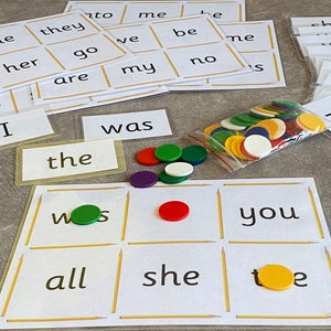 May include: A game board with nine squares, each containing a word. The words are "was", "the", "you", "all", "she", "toe", and "me". There are also colorful chips on the board and in a plastic bag.
